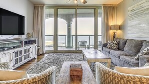 A living room with a flat-screen TV, a ceiling fan, a sofa, a coffee table, and a view of the ocean through large sliding doors. - Florida Resort Condo: Walk to Pensacola Beach! (Pensacola Beach)