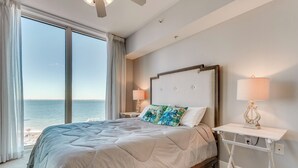 A bedroom with a large bed, a nightstand with a lamp, and a view of the ocean. - Florida Resort Condo: Walk to Pensacola Beach! (Pensacola Beach)