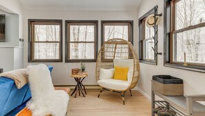 Ein gemütliches Zimmer mit einem blauen Sofa, einem weißen, flauschigen Sessel und einem Korbstuhl mit gelbem Kissen. Es gibt drei große Fenster mit Blick auf Bäume, einen kleinen Holztisch mit Büchern und einer Pflanze sowie einen Korb auf einem Regal.