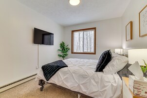 Un dormitorio con una cama, un televisor montado en la pared, una ventana con vista a árboles y una mesita de noche con una lámpara y una planta.