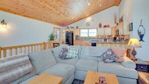 Ein gemütlicher Wohnbereich mit Holzboden, einer Couch mit gemusterten Kissen, einem Esstisch und einer Küche im Hintergrund.