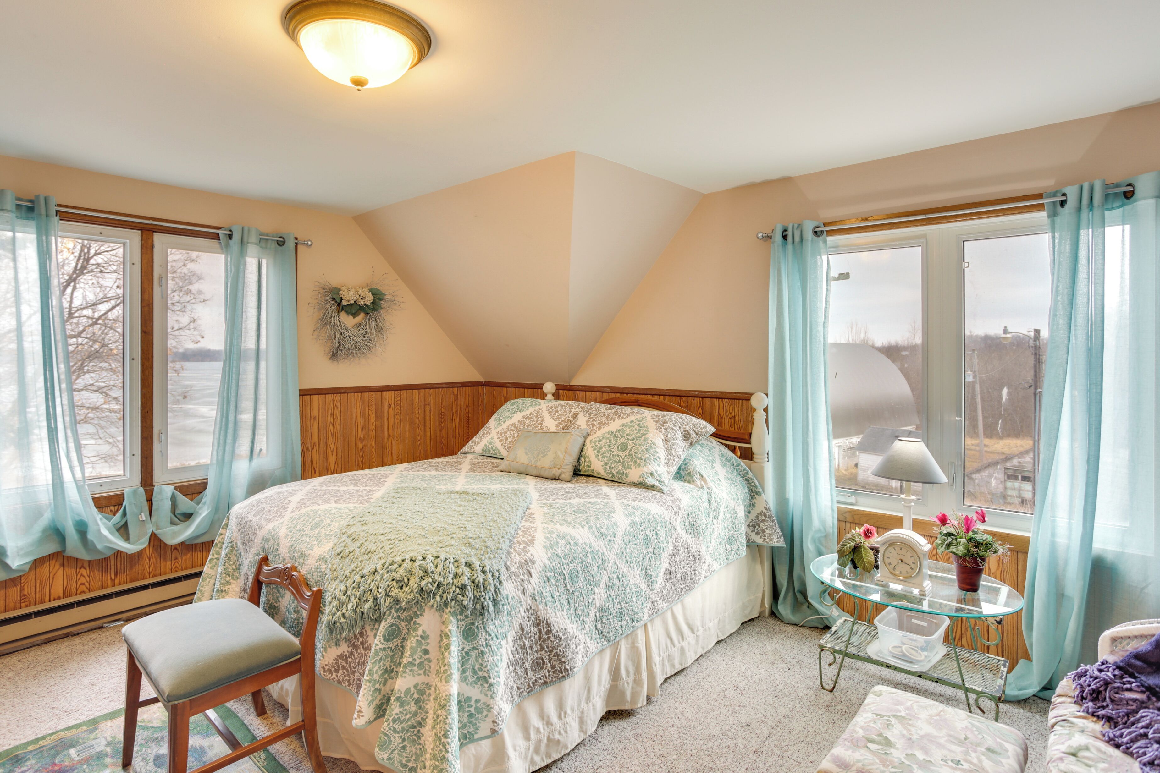 A bedroom with a bed, a chair, a bedside table with a clock and flowers, and a view of the outdoors through a window.