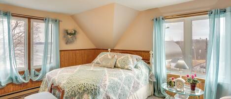 A bedroom with a bed, a chair, a bedside table with a clock and flowers, and a view of the outdoors through a window.