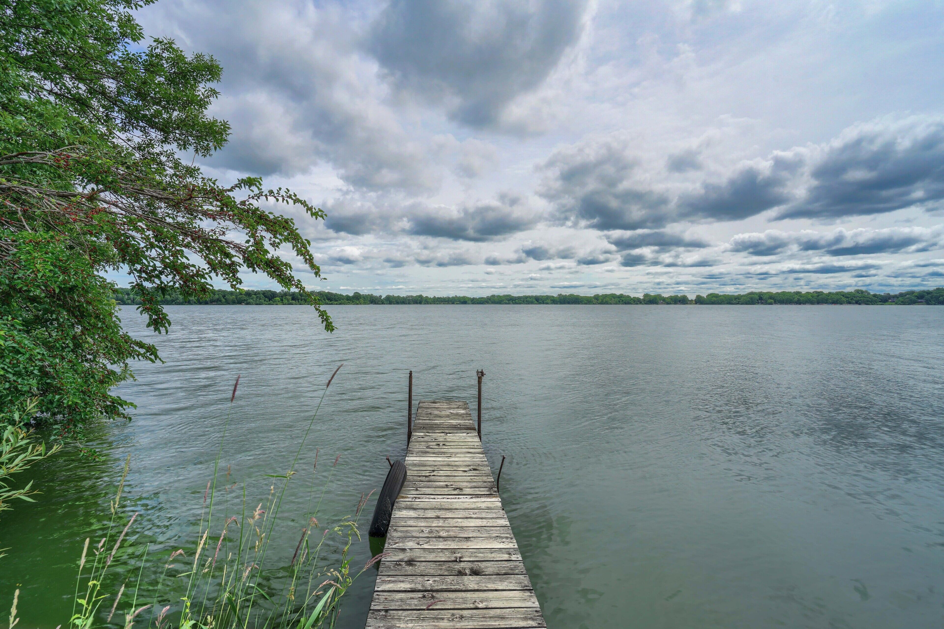 A wooden pier extending into a calm lake, surrounded by greenery and under a cloudy sky.