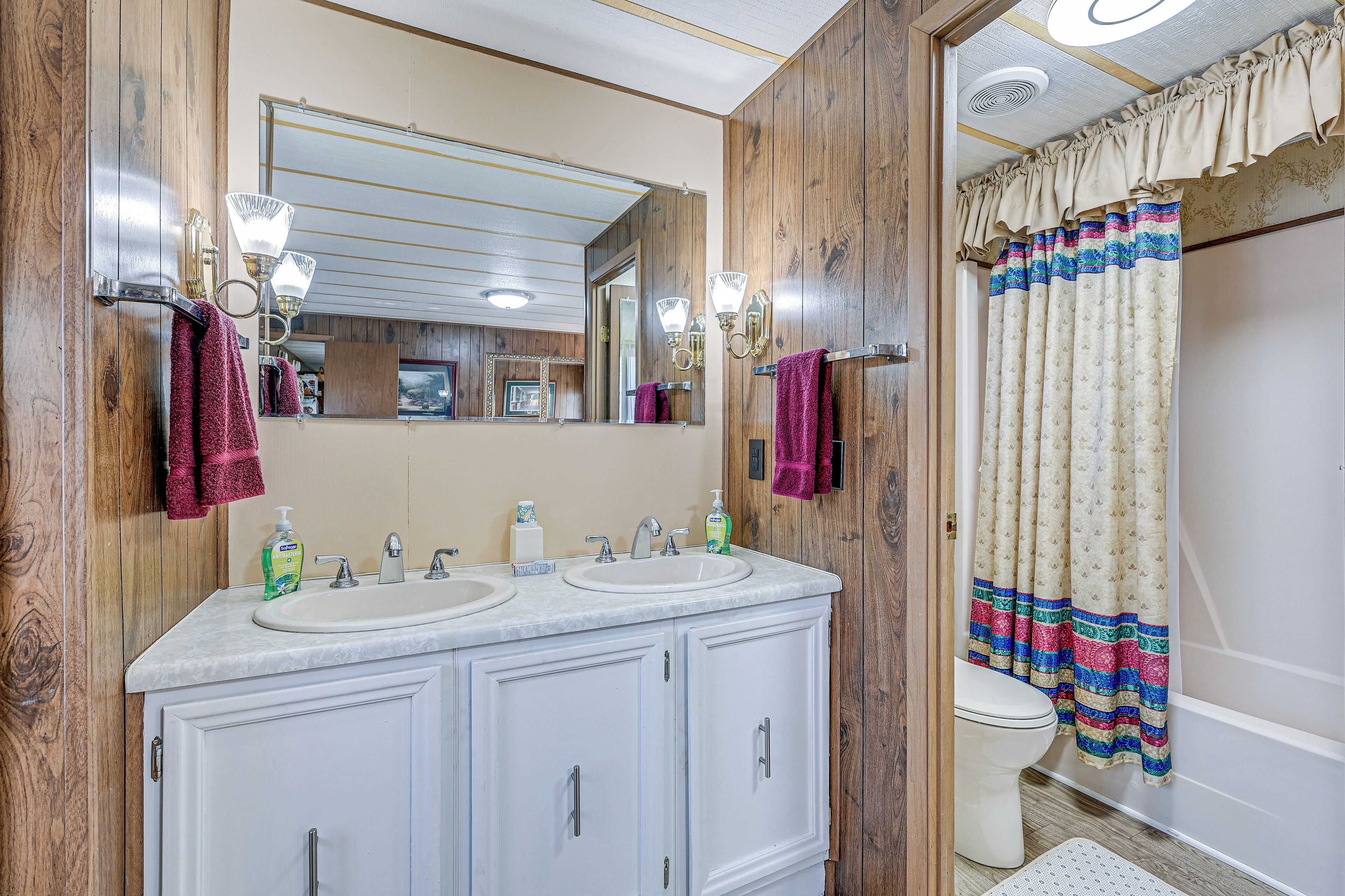 A bathroom with wooden paneling, a double sink vanity, a toilet, and a shower with a colorful curtain.