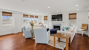 A spacious living room with hardwood flooring, a white fireplace, and a large flat-screen TV mounted above it. - Stroll to Chatham's Popular Shops & Dining / Or Walk to Andrew Hardings Beach from This NEW Impeccable Compound (Chatham)