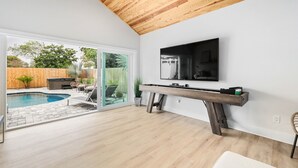Una sala de estar moderna con techo de madera, un televisor grande montado en la pared, y una piscina visible a través de puertas de vidrio, además de un banco de madera.