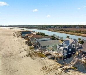 Ein direkt am Strand gelegenes Anwesen mit mehreren Gebäuden, darunter ein großes Haus mit Balkon, ein kleineres Haus und ein Bauwerk mit Treppe zum Strand.