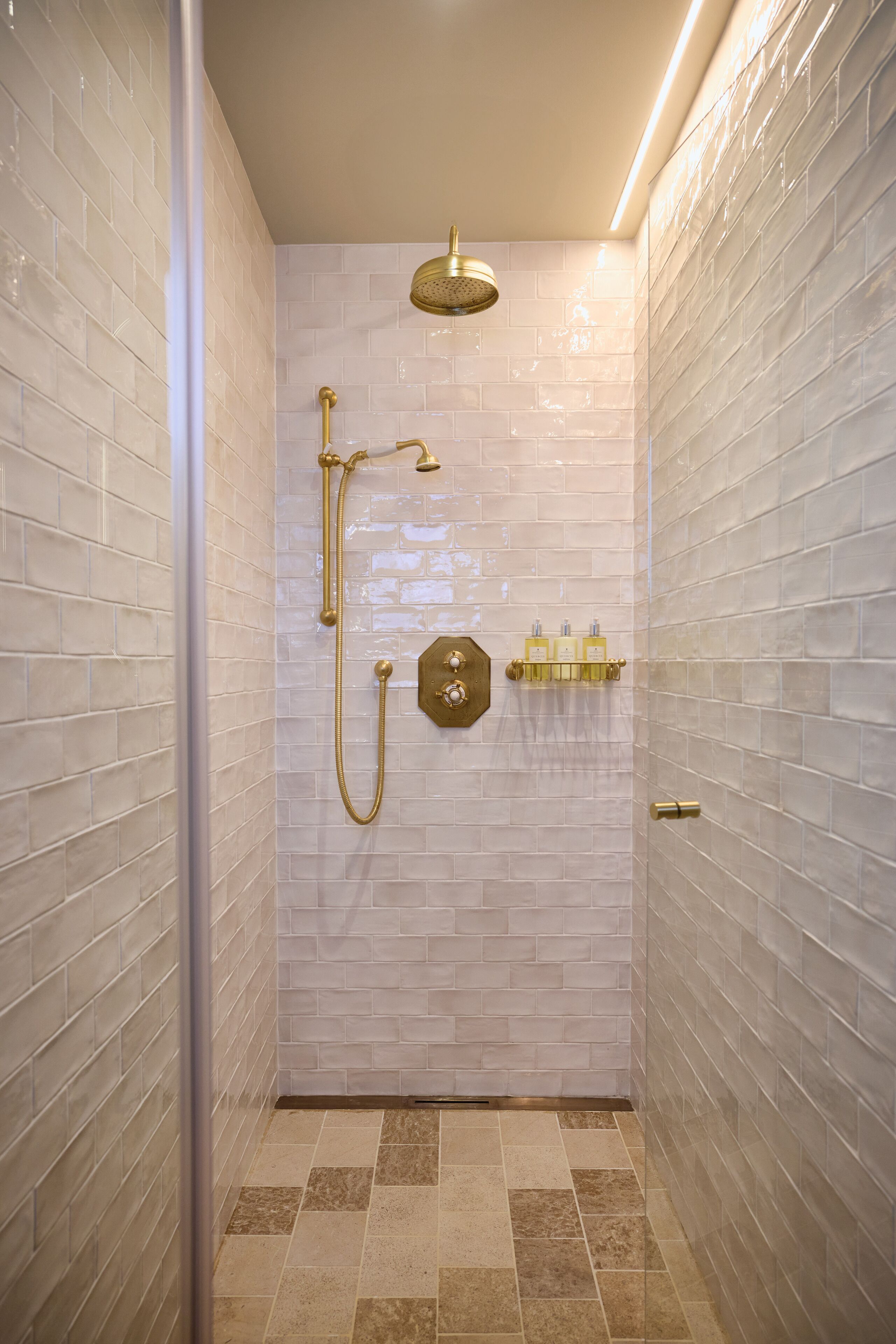 A walk-in shower with white tiled walls, a gold showerhead, and a handheld showerhead.