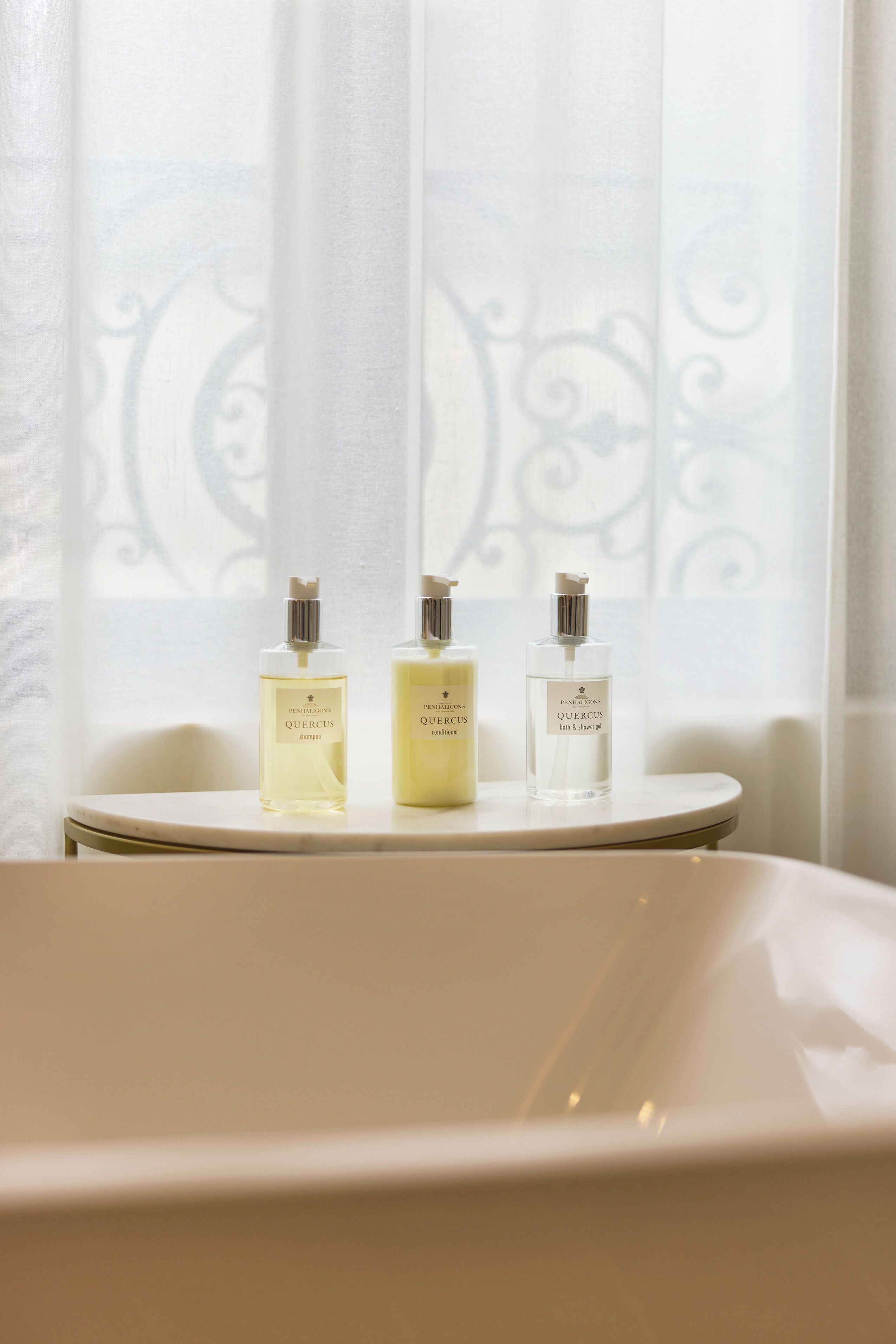 A bathroom with a bathtub and three bottles of Quercus shampoo, conditioner, and body wash on a small table.