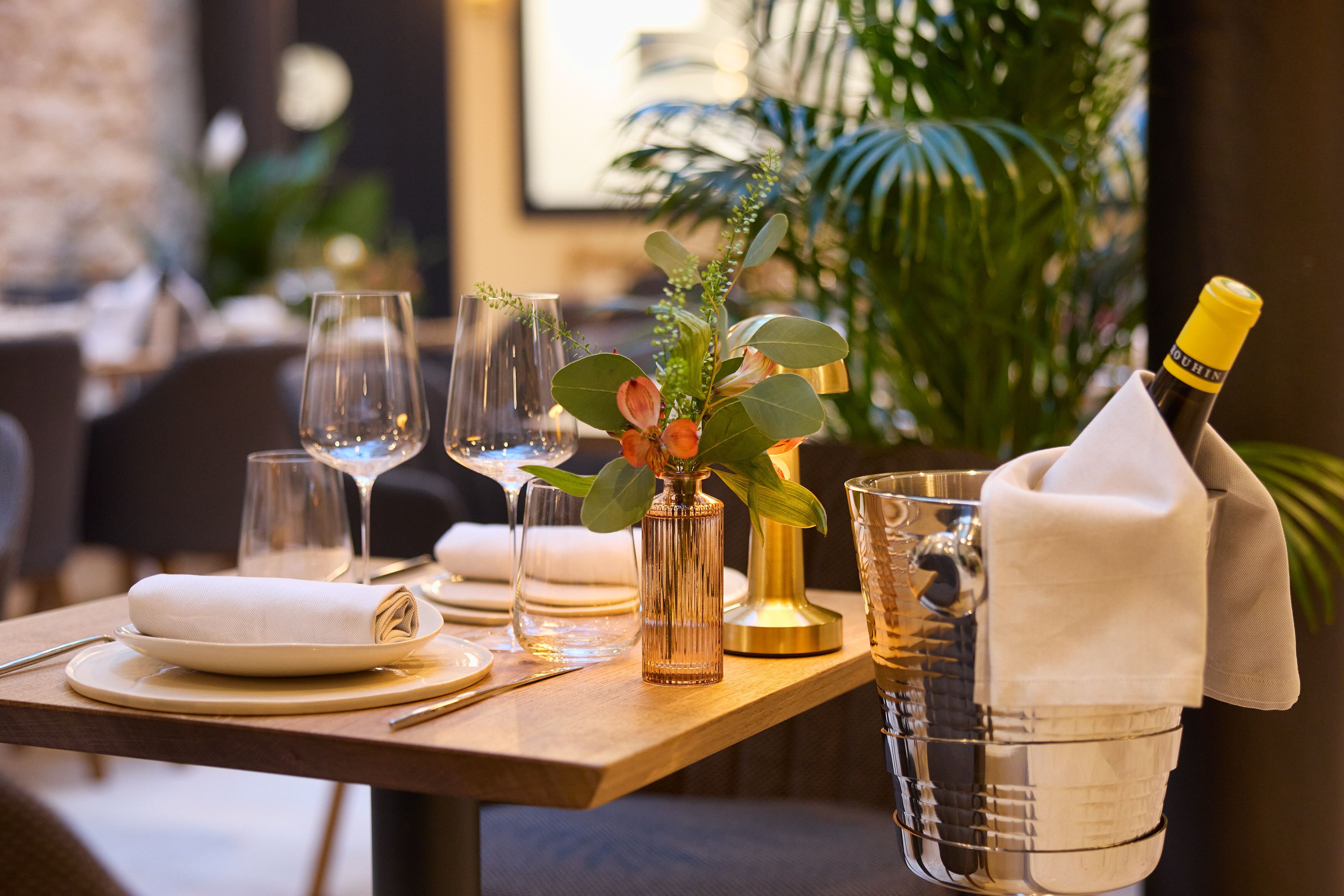 A well-set dining table with a bottle of champagne, a vase with flowers, and a glass pitcher.