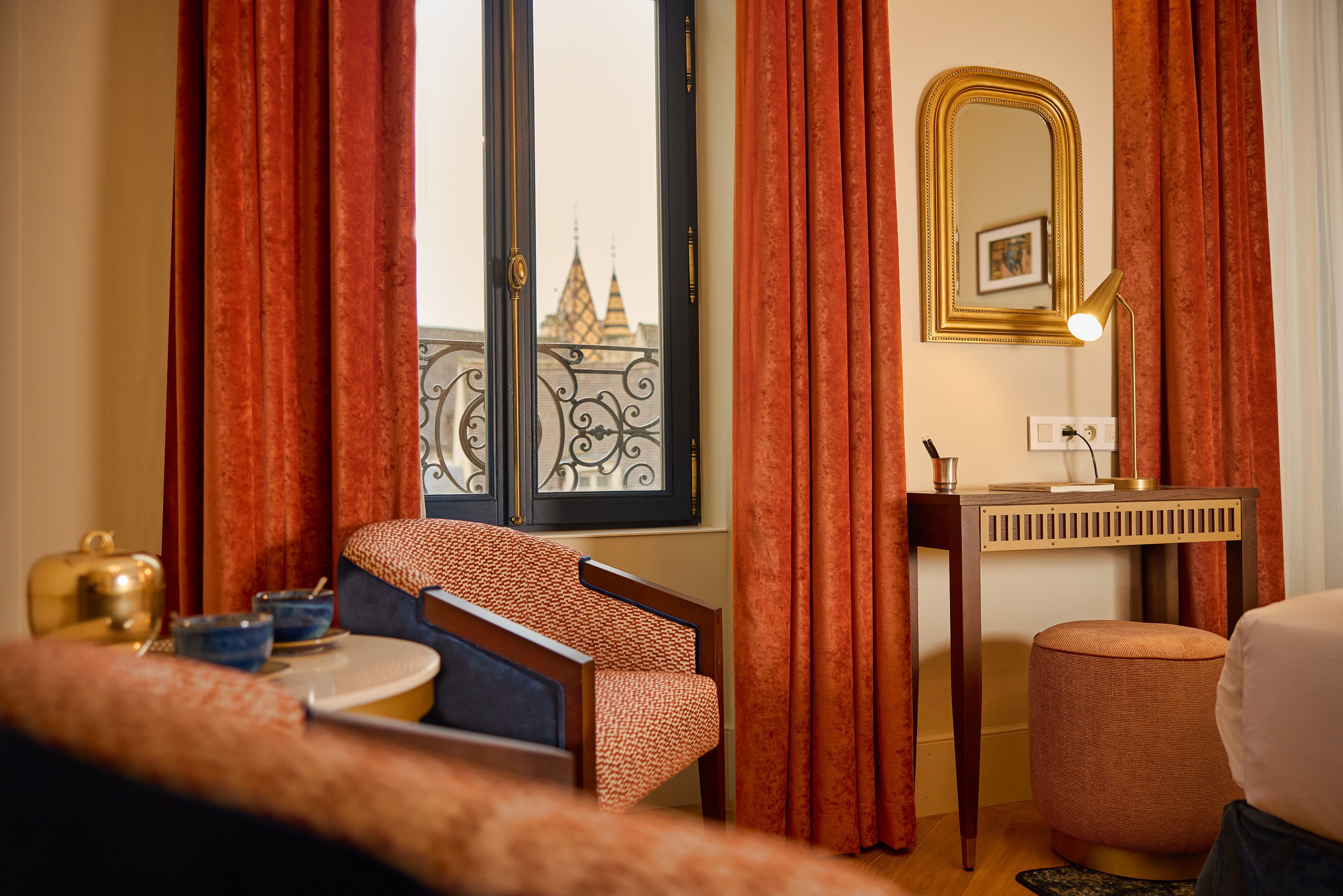 Quarto de hotel com vista para um edifício através de uma janela, uma cama, uma cadeira, uma mesinha com um bule e chá, uma escrivaninha com uma luminária e um espelho.