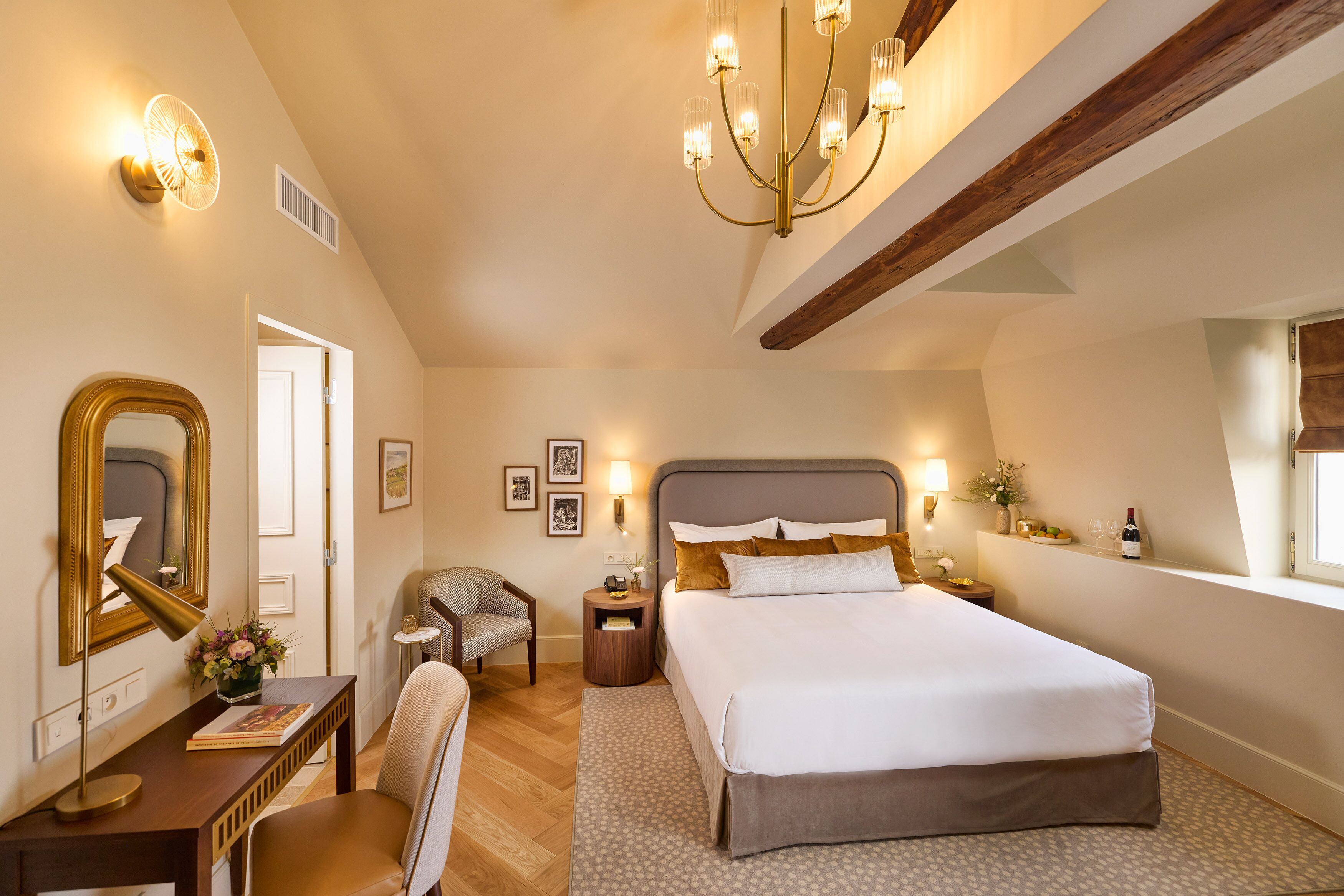A bedroom with a large bed, a desk with a chair, a mirror, and a chandelier.