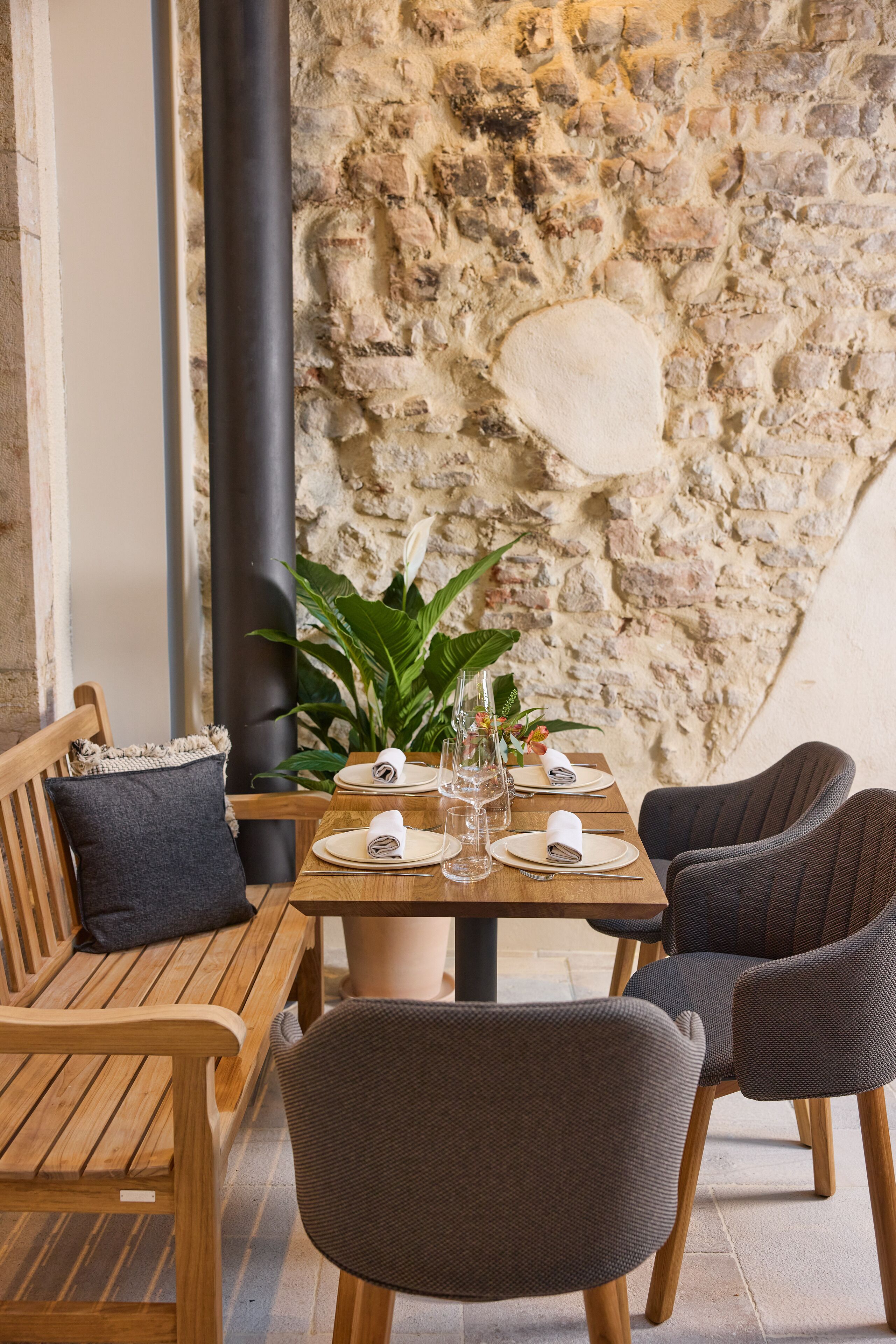 A wooden table set with plates, glasses, and a vase, against a stone wall.