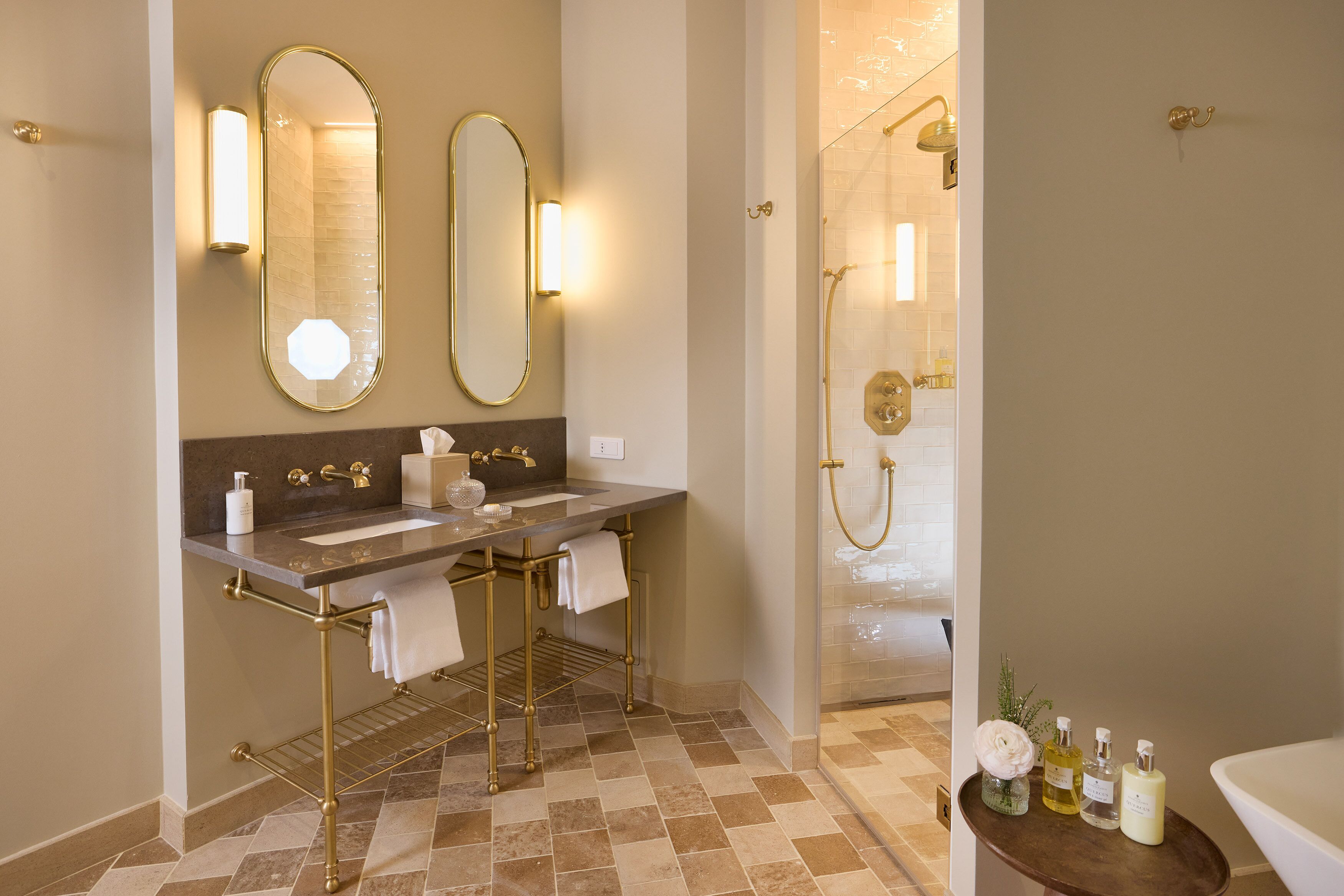 A bathroom with a double sink, two mirrors, a shower with a glass door, and toiletries on a side table.