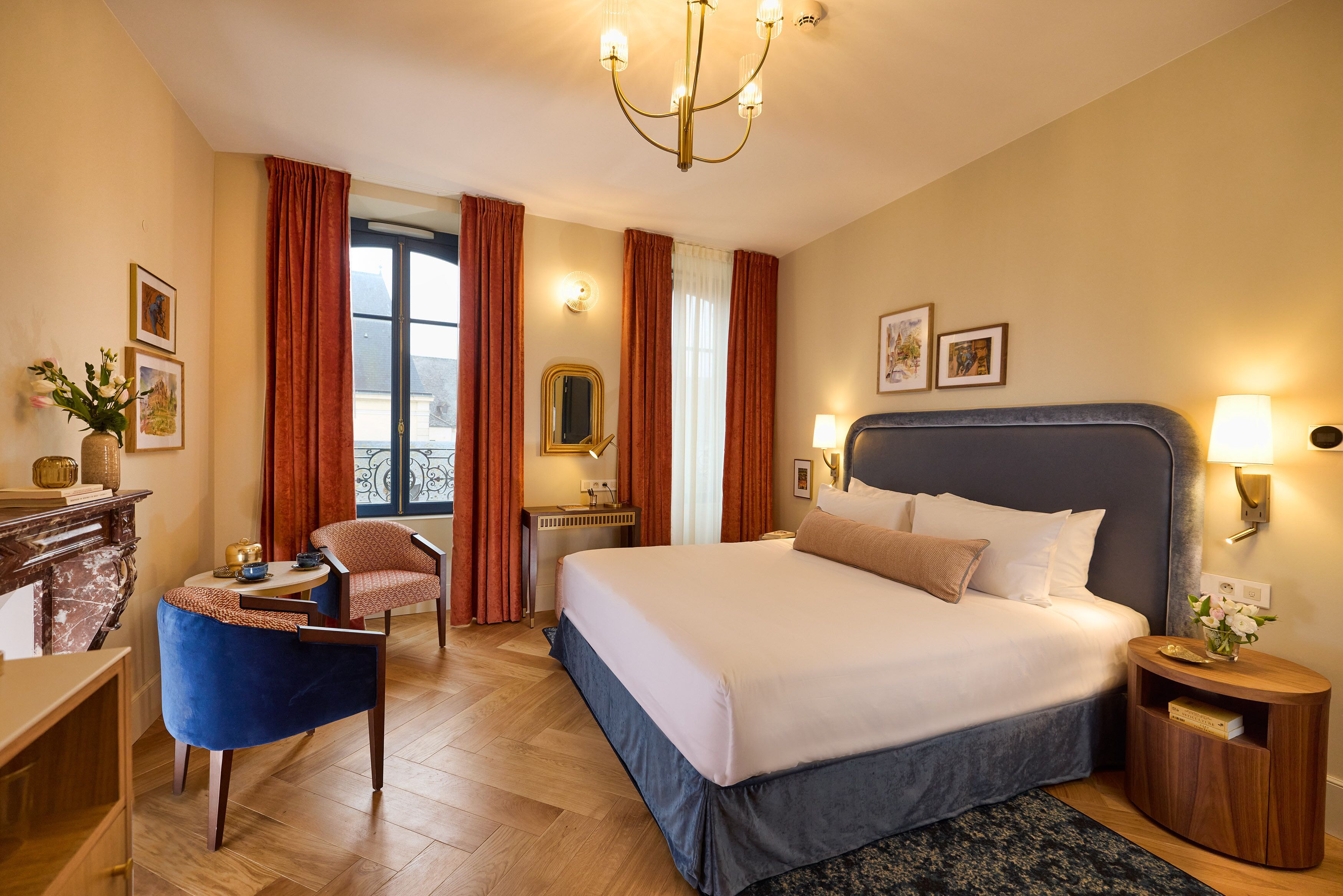 A hotel room with a large bed, a desk with a chair, a small table, a lamp, and a view of the outdoors.
