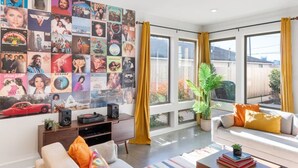 Una sala de estar moderna con una gran pared de portadas de discos, un sofá con cojines de colores, una mesa de centro con libros y un ventanal con cortinas.