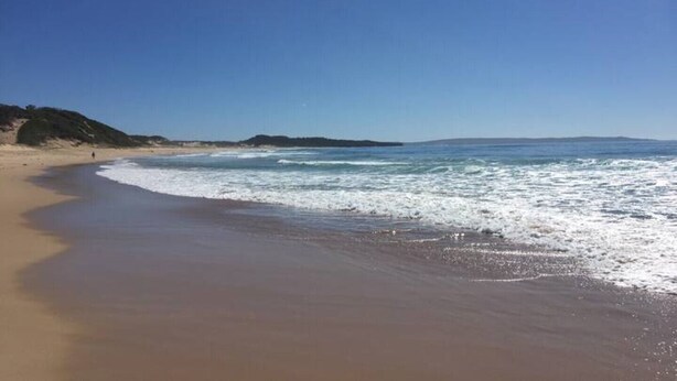 Una extensa playa de arena con olas suaves, cielo azul despejado y colinas a lo lejos.