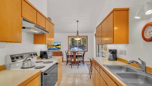 A compact kitchen with wooden cabinets, a stove, a sink, and a dining area with a table and chairs. - Single-Level 3BR | AC | River, Trails (Bend)