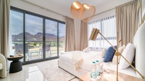 A modern bedroom with a large bed, a bedside table with a lamp, and a view of mountains through the floor-to-ceiling windows. - Beautiful 3BR Villa with Assistant Room Al Dana Island, Fujairah by Deluxe Holiday Homes (Fujairah)