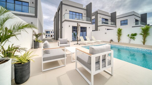A modern outdoor pool area with white furniture, potted plants, and a clear blue pool. - Beautiful 3BR Villa with Assistant Room Al Dana Island, Fujairah by Deluxe Holiday Homes (Fujairah)