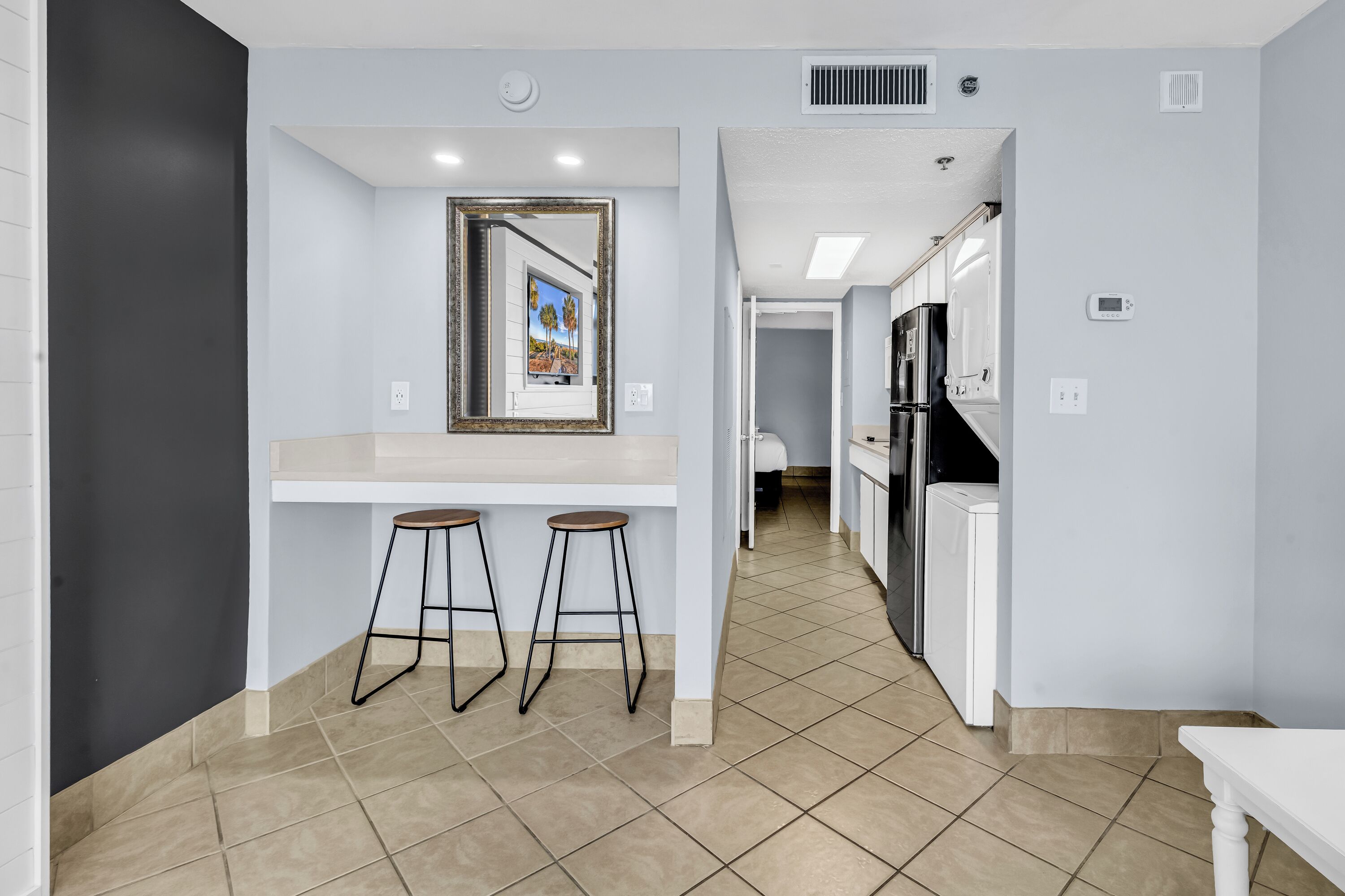A modern kitchen with a white countertop, two stools, a refrigerator, and a framed picture on the wall.