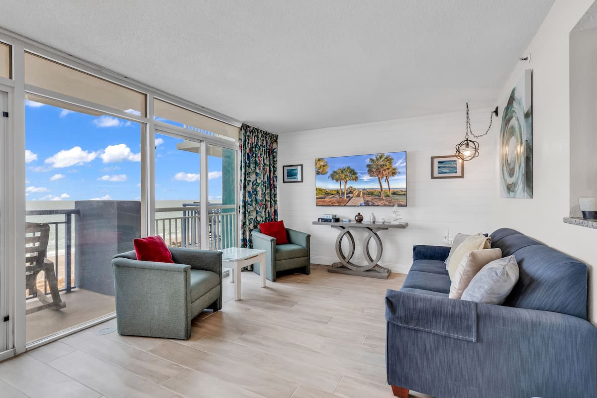A spacious living area with a balcony, a grey sofa, a small table, and a painting of a landscape.