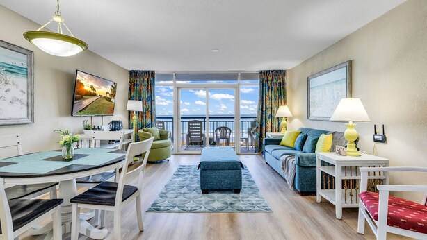 A modern living room with a dining area, a sofa set, a small table with a lamp, and a view of the sea through large windows. - Atlantica Resort Myrtle Beach (Myrtle Beach)