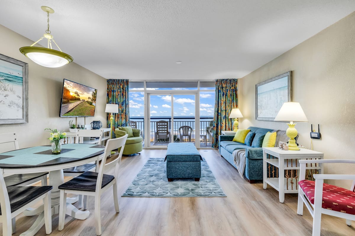 A modern living room with a dining area, a sofa set, a bench, and a view of the ocean through large windows.