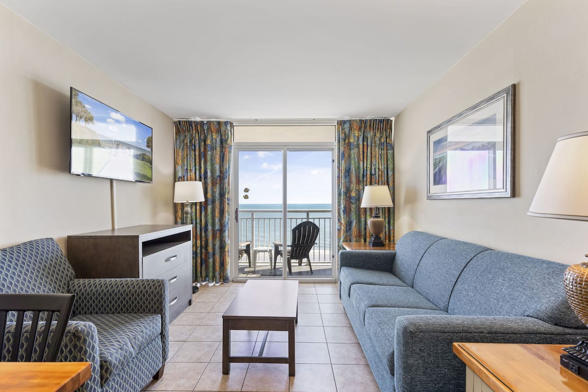 A room with a sofa, a table, a TV, and a balcony with a view.