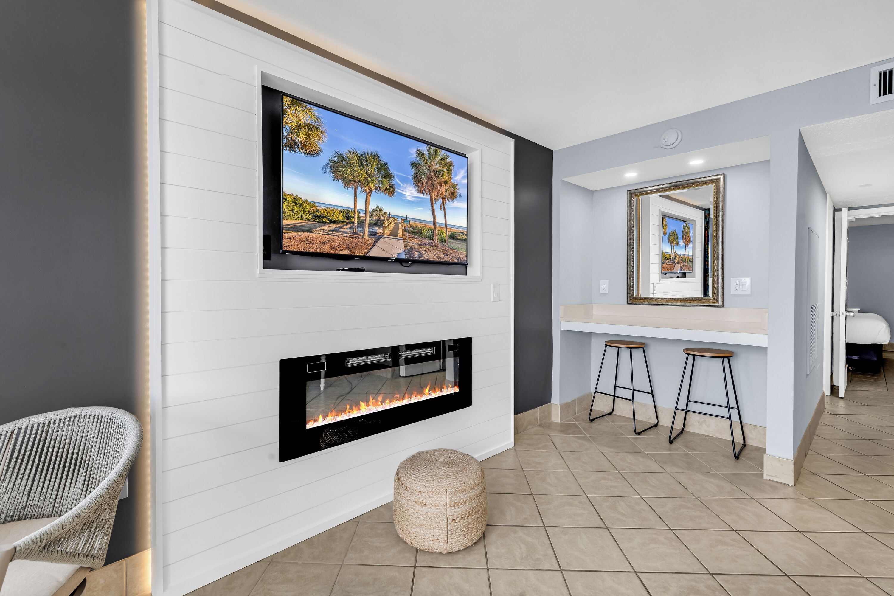 A modern living room with a flat-screen TV, a fireplace, a woven ottoman, and a bar area with stools.