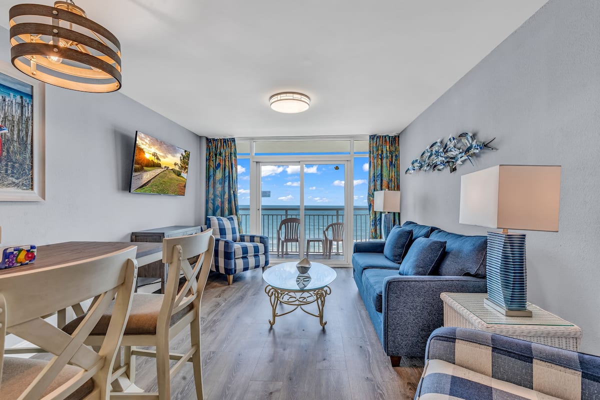 A modern living room with a dining area, a sofa set, a coffee table, and a view of the ocean through large windows.