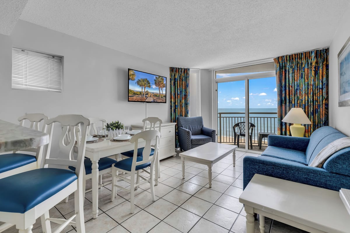 A modern living room with a dining area, a sofa, a TV, and a balcony with a view.
