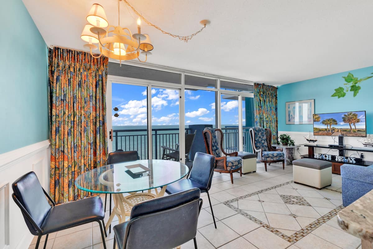 A spacious living area with a glass table, comfortable seating, a balcony with a view, and a beach-themed decor.