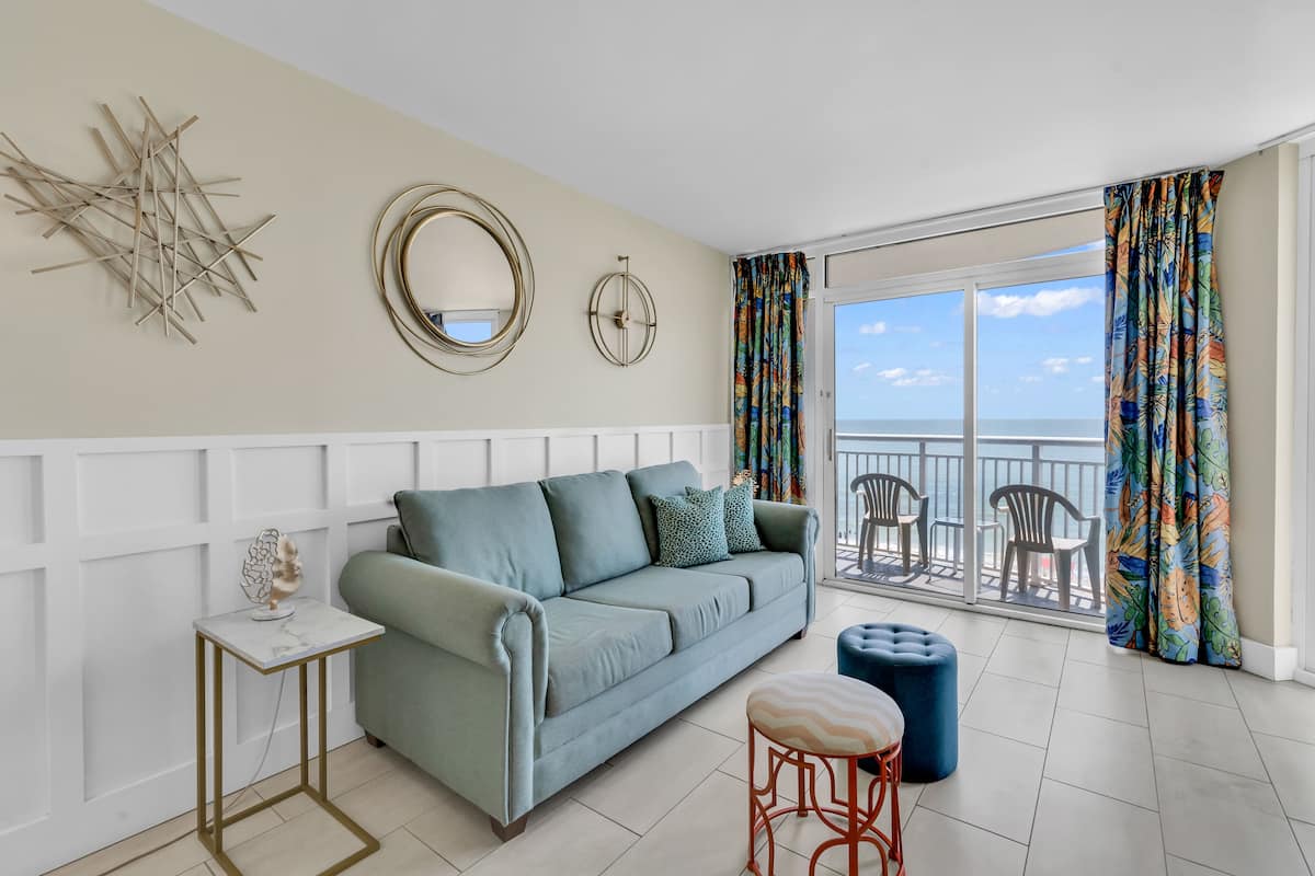 A living room with a sofa, a side table, a small stool, and a balcony with chairs.