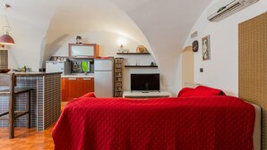 A compact room with a kitchenette, a bed with a red blanket, a television, and a small dining area.