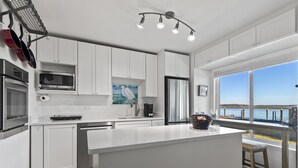 Une cuisine moderne avec des meubles blancs, des appareils en acier inoxydable, un grand îlot central et une vue sur le bord de l’eau.