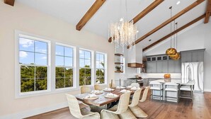 A spacious dining area with a long wooden table and wicker chairs, large windows with a view of greenery, and a modern kitchen with grey cabinets. - New, Loaded! 6-Seater LSV, 6 Bikes, Private Pool, Walk to Beach! (Santa Rosa Beach)