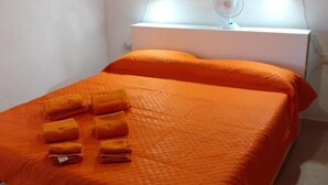Cama bem arrumada com colcha e almofadas laranja, mesa de cabeceira branca com abajur e um pequeno ventilador.