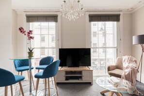 A living room with a TV, blue chairs, a glass coffee table, a sofa, and a chandelier. - Two Bedroom Apartment in the Centre of Cheltenham (Gloucestershire)