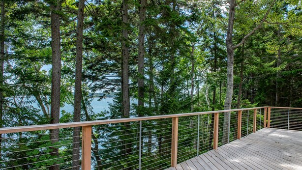 A wooden deck with a metal railing overlooking a forested area and a body of water. - Coastal Hideaway. Water views with expansive deck, pool table, office area. (Harpswell)