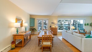 A spacious living area with a wooden dining table, a sofa, and a large window offering a view of trees. - Coastal Hideaway. Water views with expansive deck, pool table, office area. (Harpswell)
