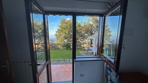 A room with a large sliding glass door leading to an outdoor area with a view of trees and a distant body of water. - AMORE RENTALS - Villa Savoia with Sea View and Garden (Sorrento)