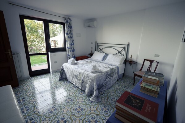 Ein Schlafzimmer mit einem Bett, einem Sessel, einem Tisch mit Büchern und einem Fenster mit Vorhängen.