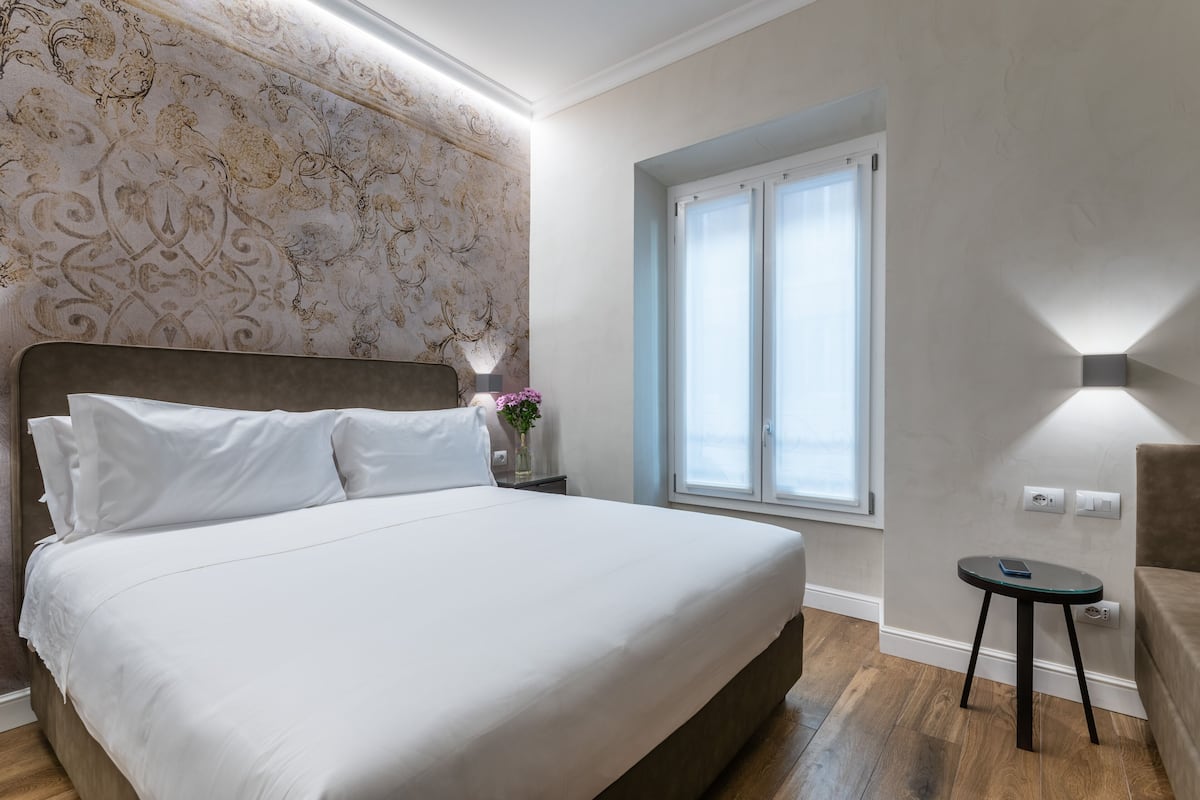 Un lit bien fait, avec du linge de lit blanc, une table de chevet ornée d’un vase de fleurs, une petite table avec une lampe et une fenêtre avec des stores.