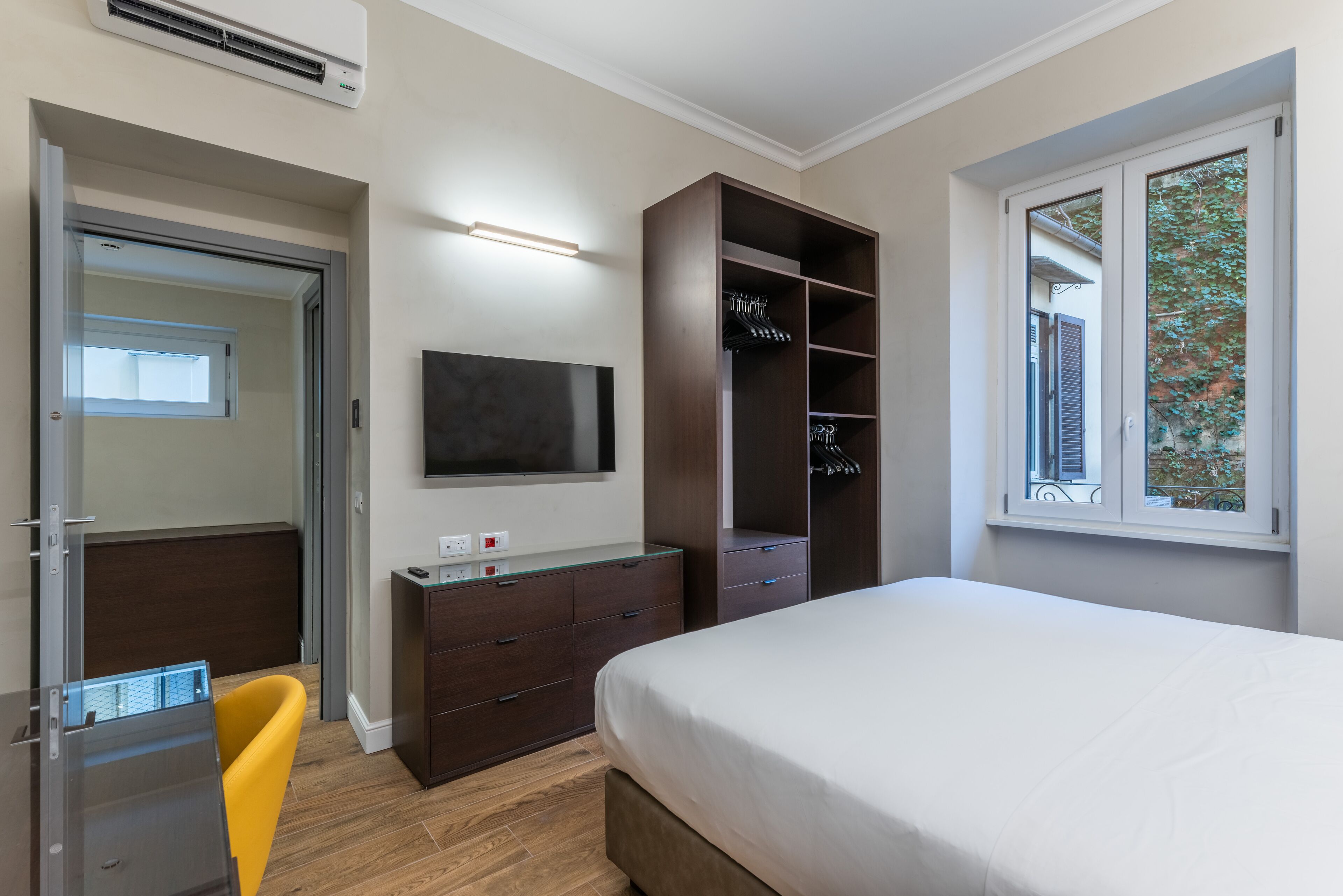 A bedroom with a bed, a TV mounted on the wall, a wardrobe, a desk with a chair, and a window with a view of greenery.