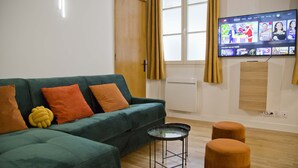 Uma sala de estar moderna com um sofá verde, almofadas laranja, uma mesa de centro de vidro e uma TV de tela plana na parede.