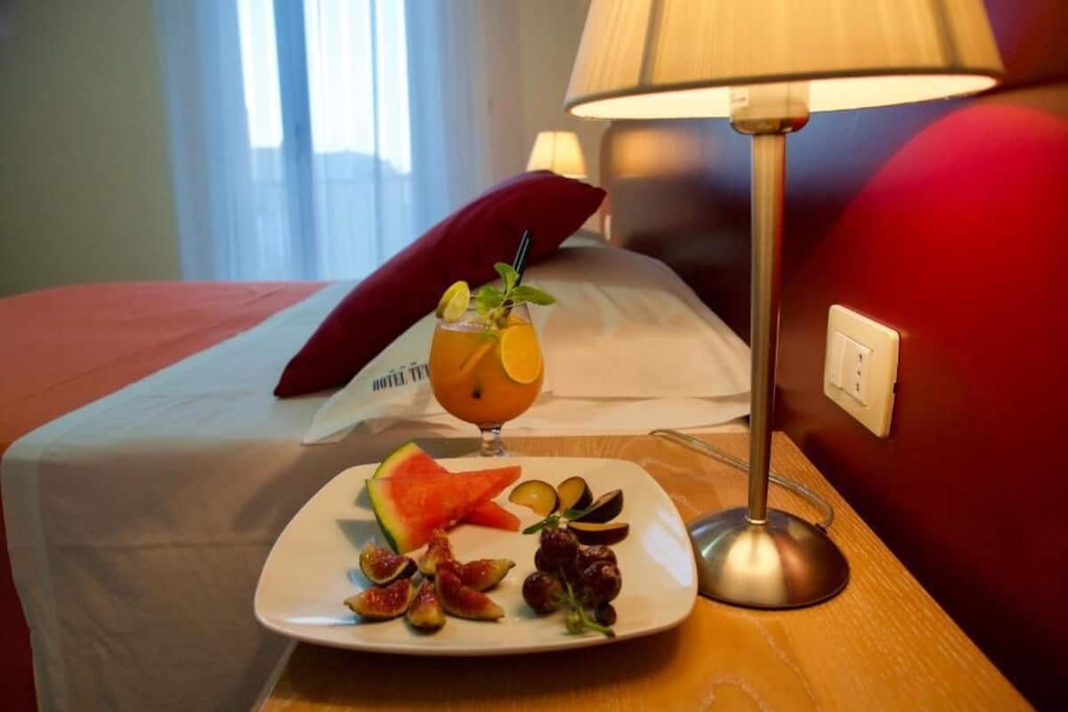 A neatly made bed with a red pillow, a plate of fruit and a glass of drink on a bedside table, and a lamp.