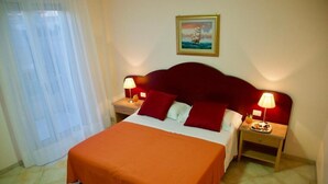 A hotel room with a bed, two bedside tables, a nightstand, a window with curtains, and a wall-mounted painting. - Hotel Teti (Ugento)