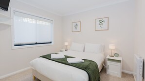 A neatly arranged bedroom with a bed, bedside tables, a window with blinds, and two framed pictures on the wall.